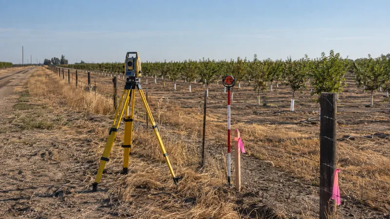 Total station on a tripod and a prism pole at a flagged stake along a wire fence line beside a young orchard